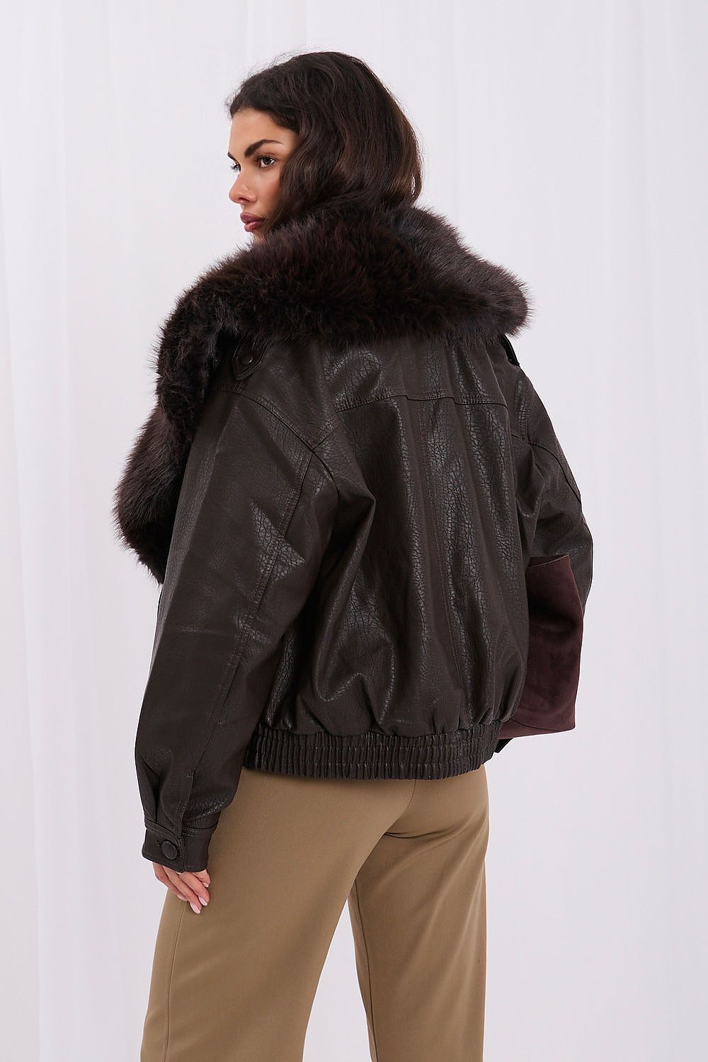 A woman shown from the back wearing a dark brown faux-leather jacket with a large dark brown fur collar and tan pants, photographed against a white studio background.