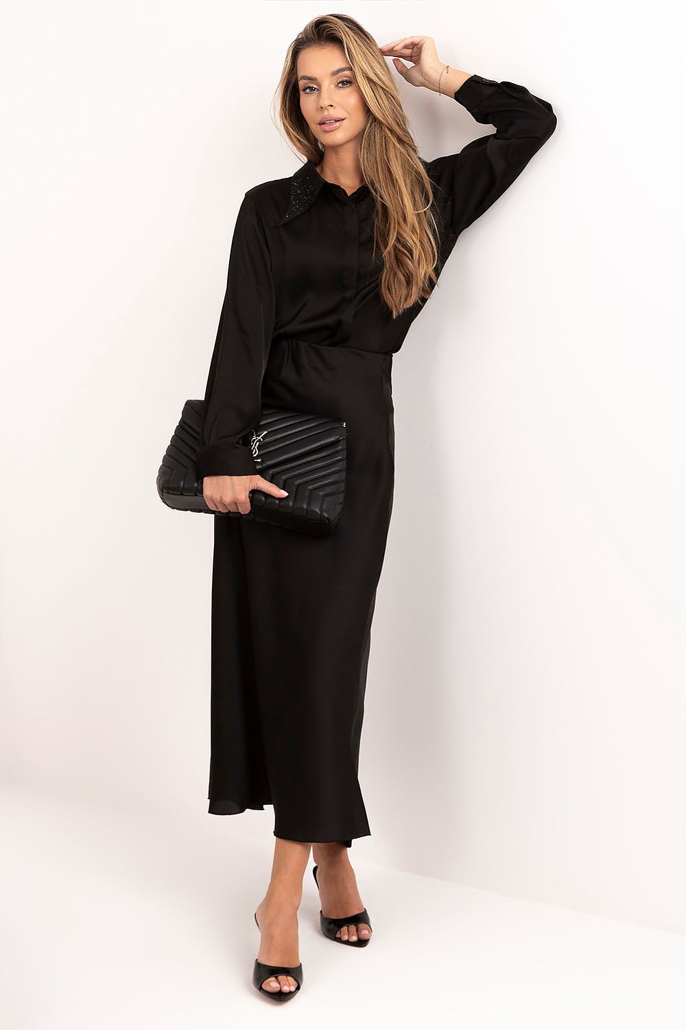 A woman modeling a black long-sleeve midi shirt dress with a collar, holding a black quilted clutch, against a plain white studio background.