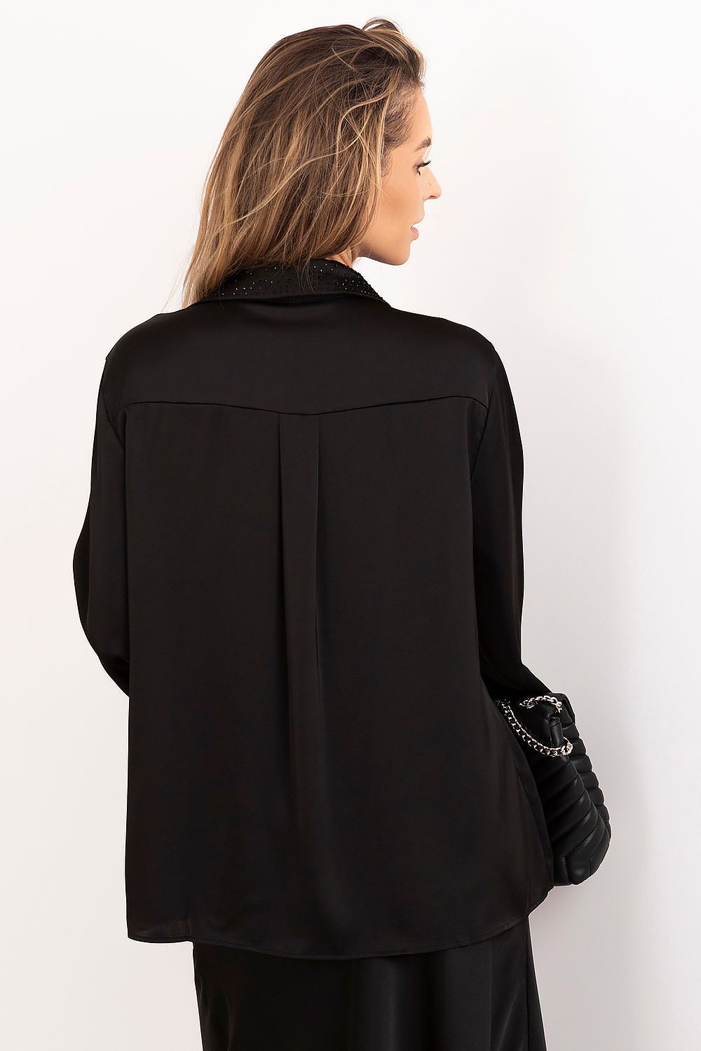 A woman shown from the back wearing a black long-sleeve blouse with a textured lace-trimmed collar and a pleated back detail, paired with black bottoms and holding a black quilted handbag.
