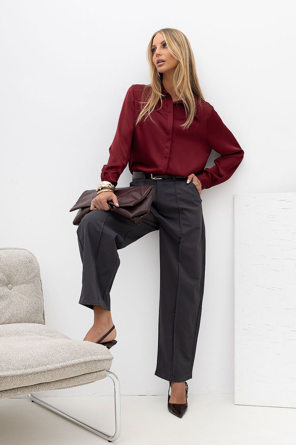 A woman models a burgundy long-sleeve satin blouse and dark charcoal gray wide-leg trousers, holding a dark brown clutch. She is posed in a minimalist studio setting with a light gray upholstered chair on the left and a white wall panel on the right.