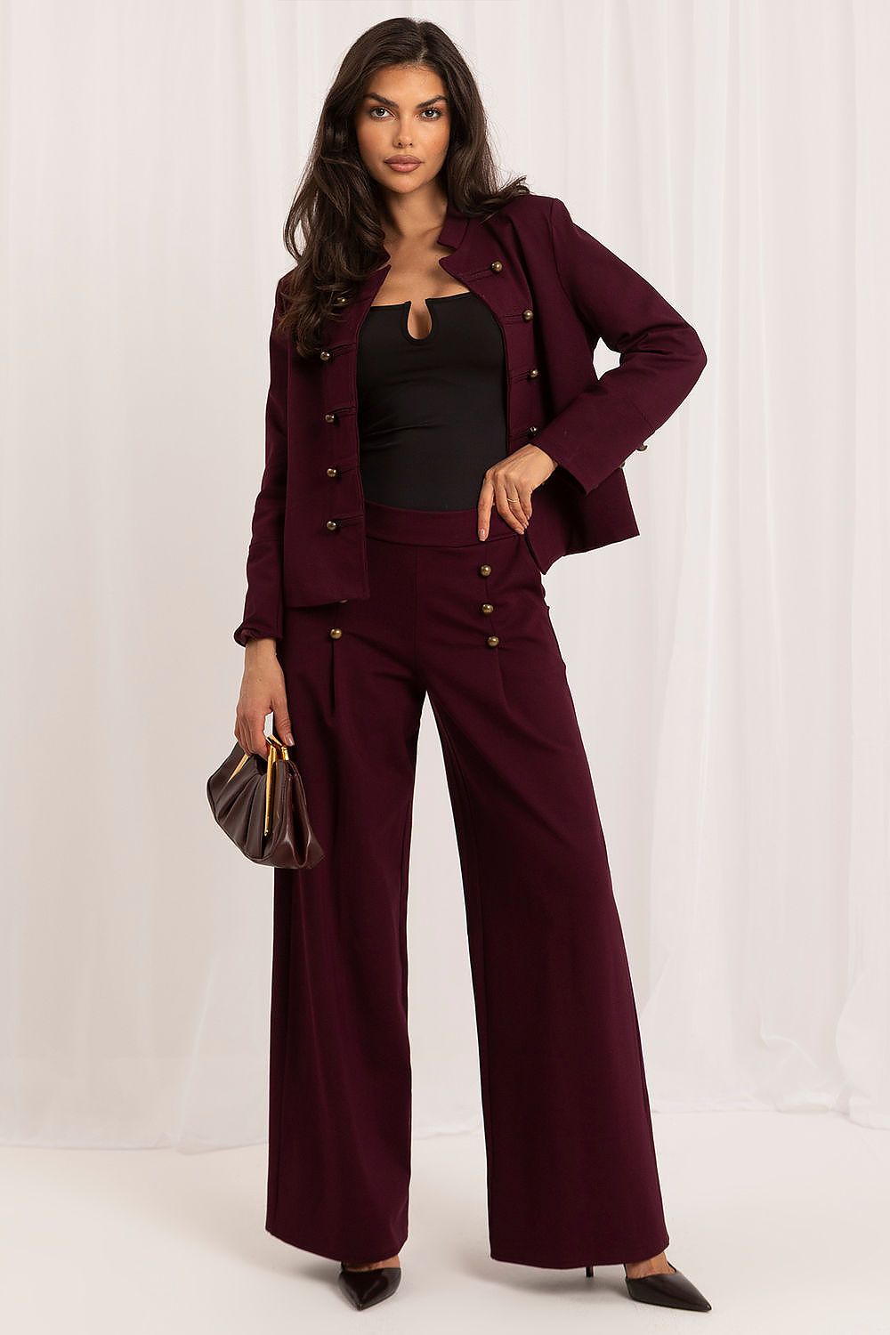 A woman models a matching burgundy two-piece suit set featuring a double-breasted blazer and wide-leg trousers with gold-tone buttons, styled with a black top, black heels, and a small pleated brown handbag against a white draped backdrop.