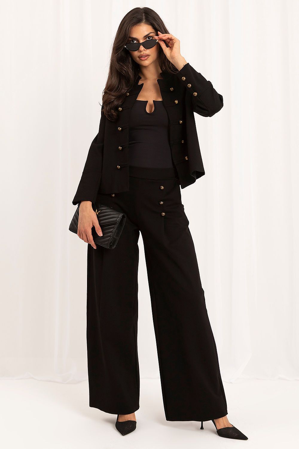 A woman modeling a black two-piece outfit consisting of a cropped jacket with gold-tone buttons and matching wide-leg pants, styled with a black top, black sunglasses, and a black clutch bag against a white draped background.