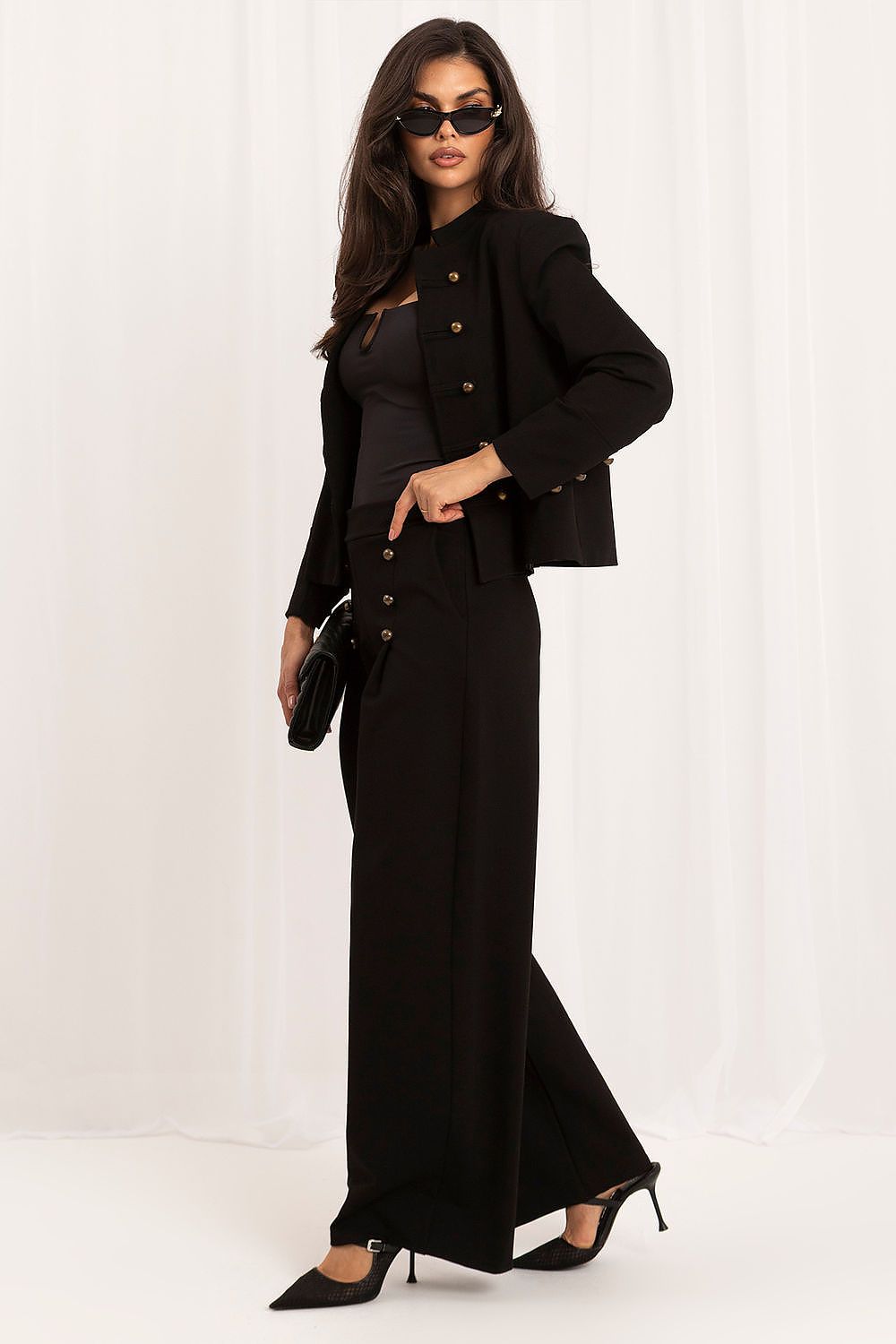 A model wearing a black two-piece outfit consisting of a cropped jacket with gold buttons and matching wide-leg trousers, styled with black sunglasses, a black clutch, and black high heels against a white draped background.