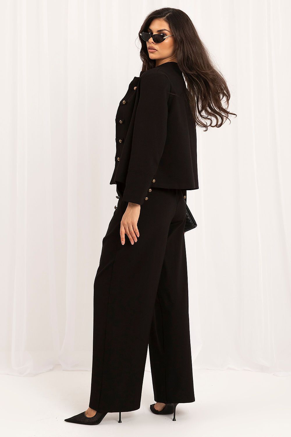 A woman modeling a black two-piece outfit consisting of a cropped jacket with gold-tone buttons and matching wide-leg trousers, photographed against a white draped fabric backdrop.