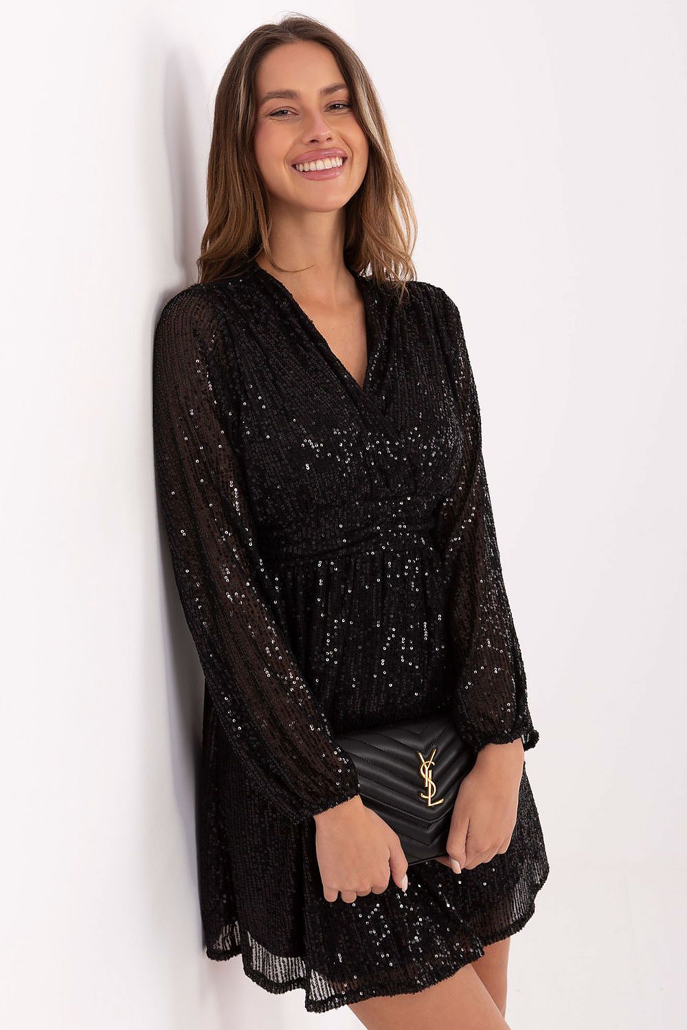 A woman wearing a black long-sleeve lace mini dress with sequins, holding a black quilted clutch with a gold YSL logo, posing against a white wall.