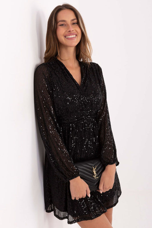 A woman wearing a black long-sleeve lace mini dress with sequins, holding a black quilted clutch with a gold YSL logo, posing against a white wall.
