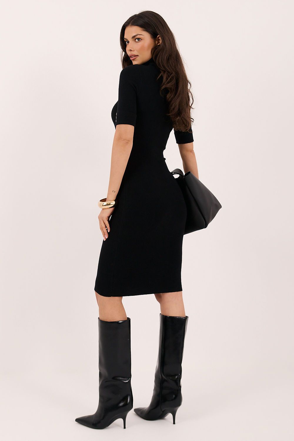 A woman modeling a fitted black short-sleeve midi dress from the back, paired with black knee-high heeled boots and a black handbag, against a plain light background.