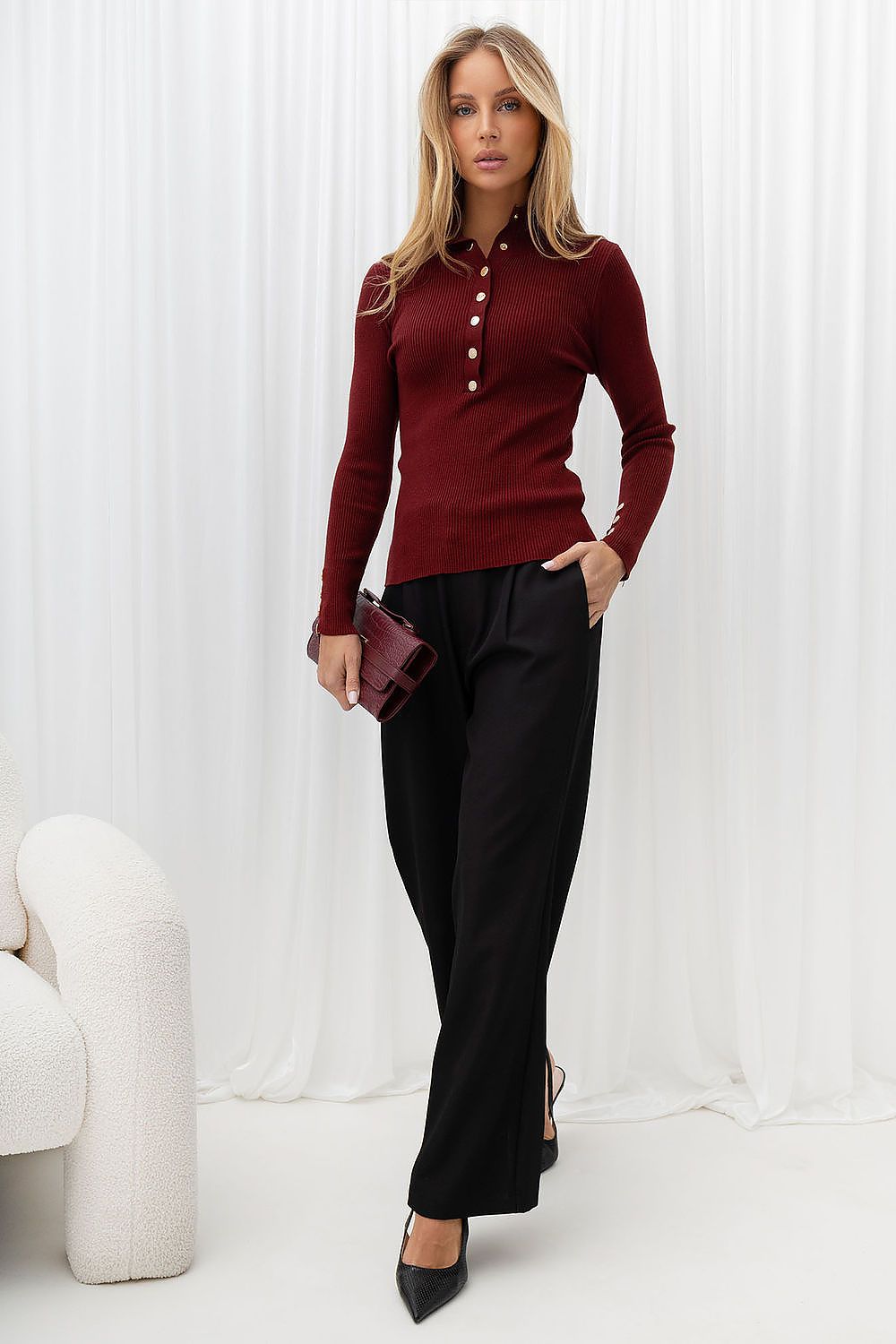 A woman models a long-sleeve burgundy knit polo shirt with a button placket, paired with black trousers and black pointed-toe heels, holding a matching burgundy clutch against a white curtain backdrop.