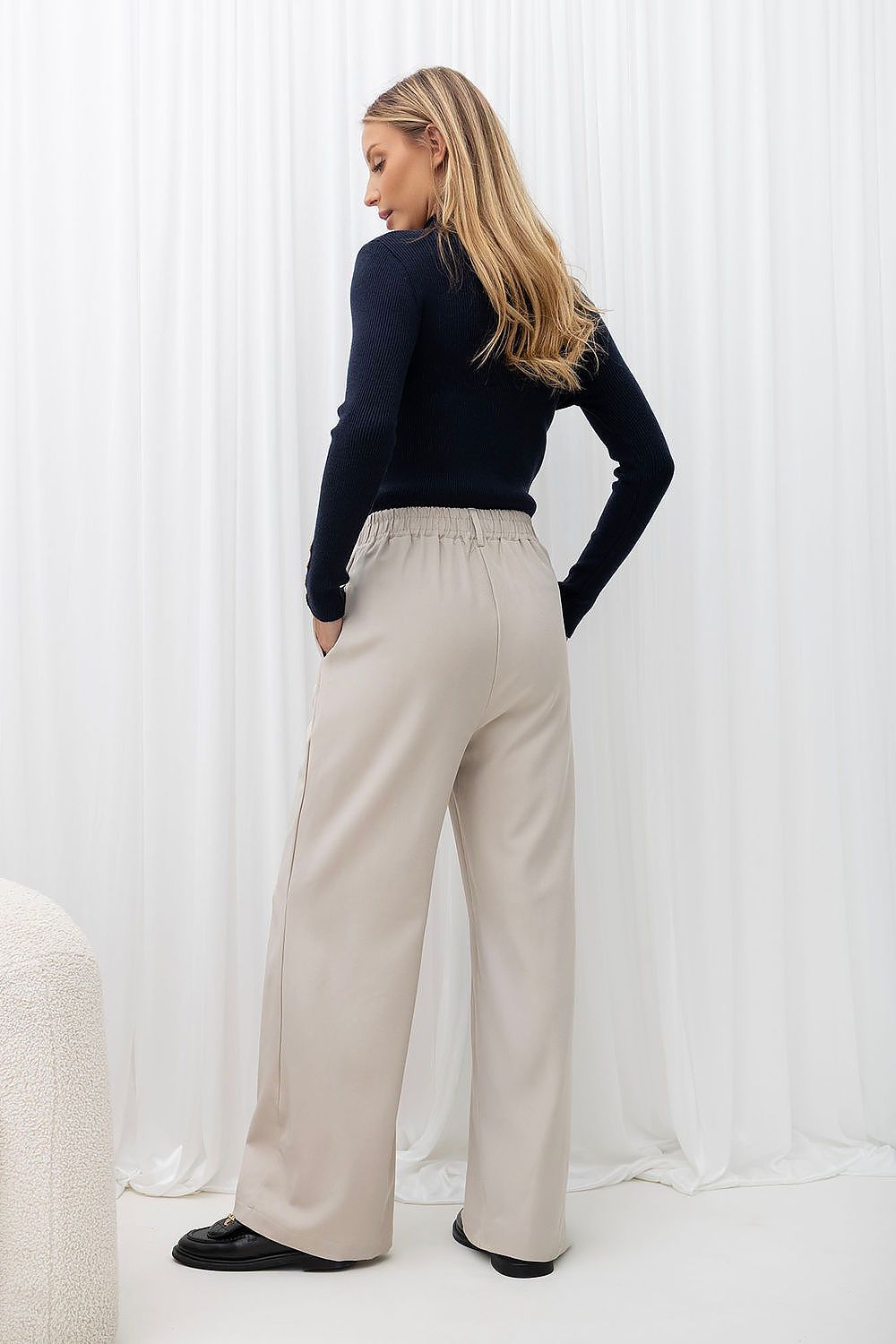 A woman stands with her back to the camera wearing high-waisted, wide-leg beige trousers, a dark navy long-sleeve top, and black shoes, posed in front of a white curtain backdrop.