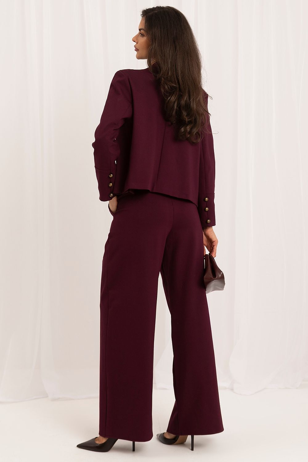 A woman modeling a matching burgundy two-piece suit with a cropped jacket and wide-leg trousers, shown from the back against a white draped studio backdrop.