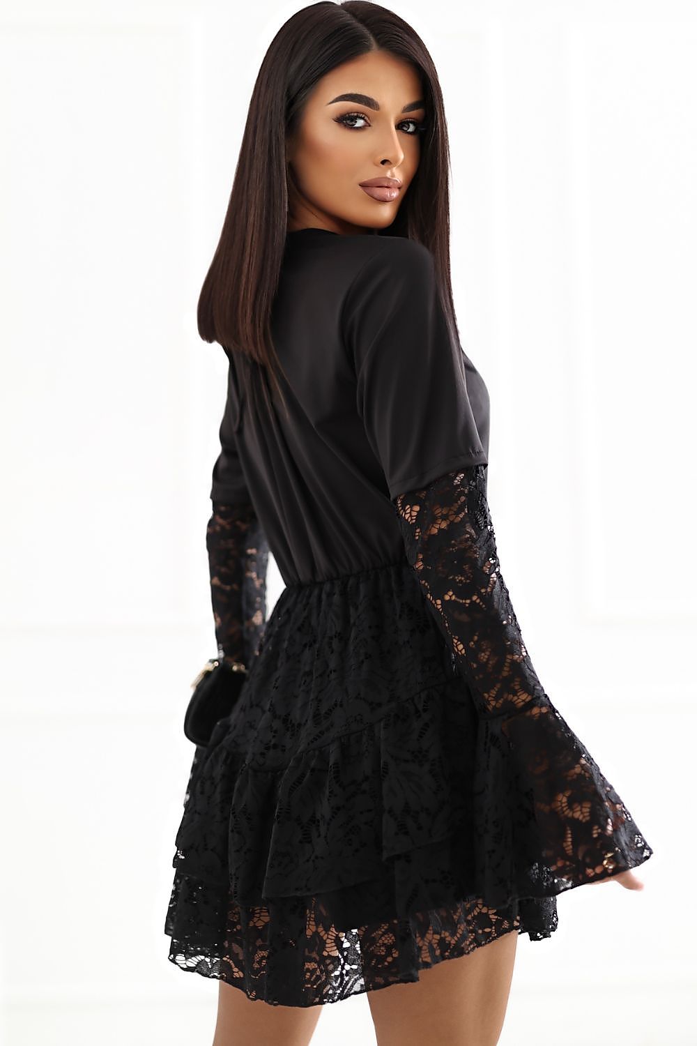 A woman modeling a black long-sleeve mini dress with sheer lace sleeves and a ruffled lace hem, shown from a three-quarter back angle against a bright white background.