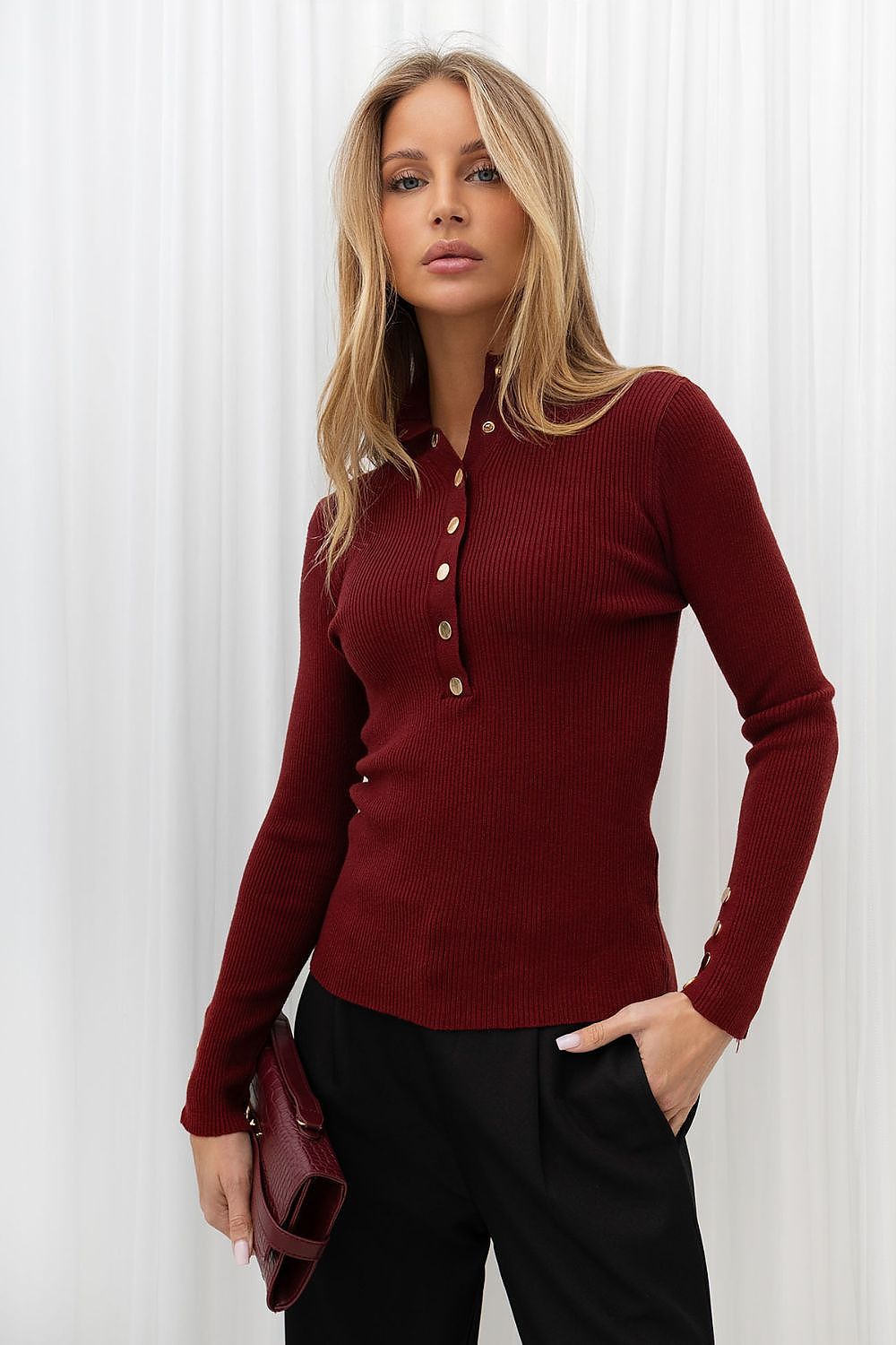 A woman modeling a ribbed long-sleeve button-front top in deep burgundy, paired with black pants, standing against a white curtain backdrop.