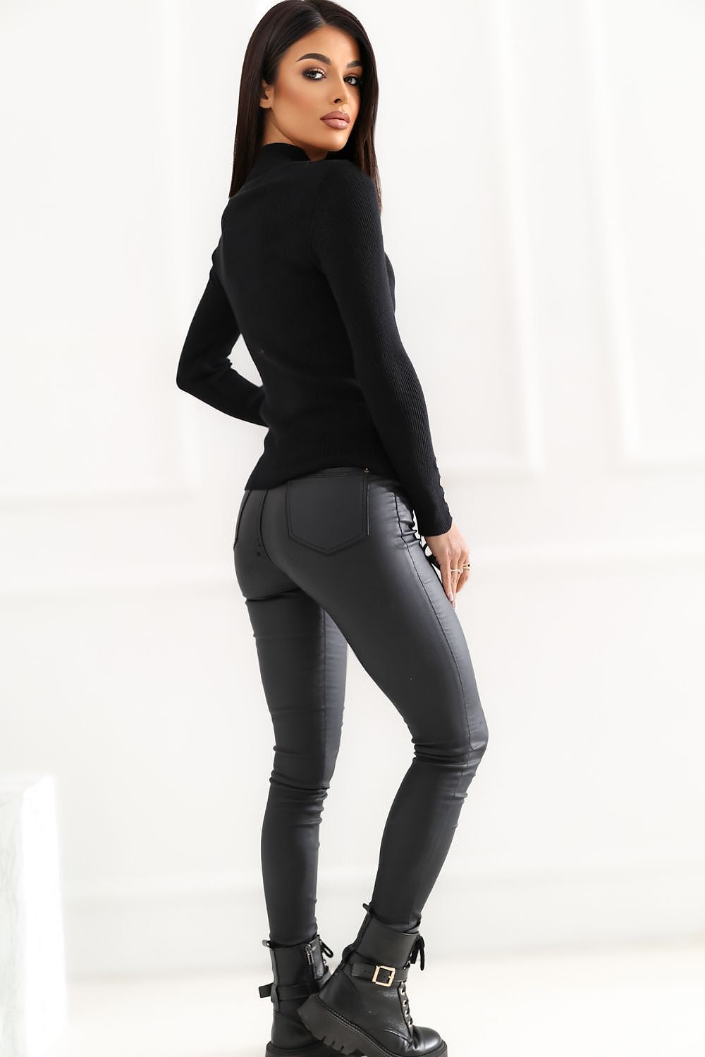 A woman posing from the back wearing a black long-sleeve mock neck top, dark gray slim-fit jeans, and black combat boots against a bright white studio background.