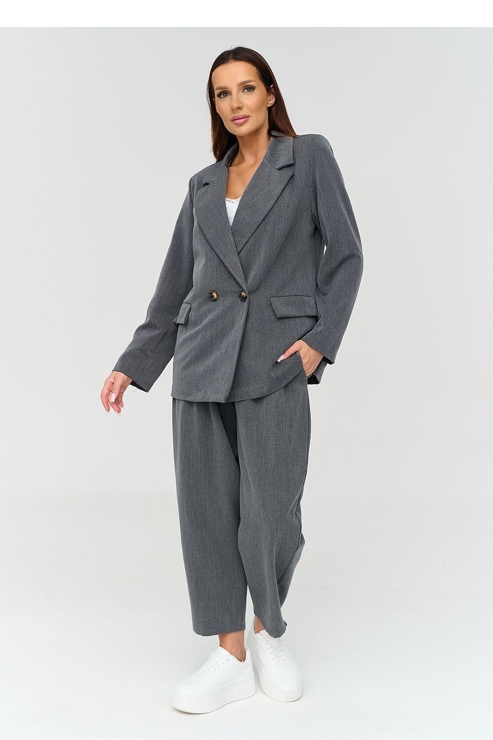 A woman modeling a gray double-breasted blazer and matching wide-leg cropped trousers, styled with a white top and white sneakers against a light gray studio background.