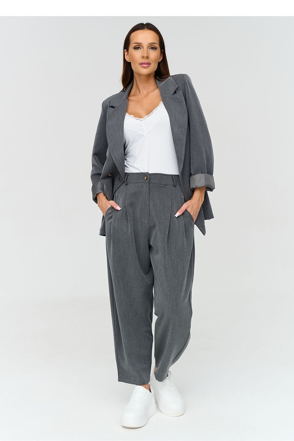 A woman modeling a gray two-piece suit set consisting of a blazer and matching wide-leg trousers, styled with a white camisole and white sneakers against a light gray studio background.