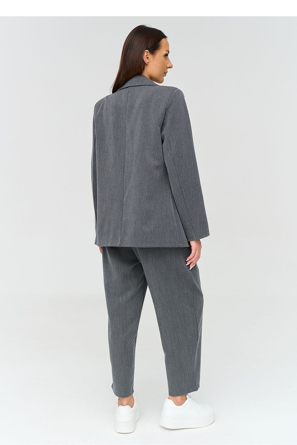 A woman shown from the back wearing a gray two-piece suit with a blazer and cropped trousers, paired with white sneakers, against a light gray studio background.