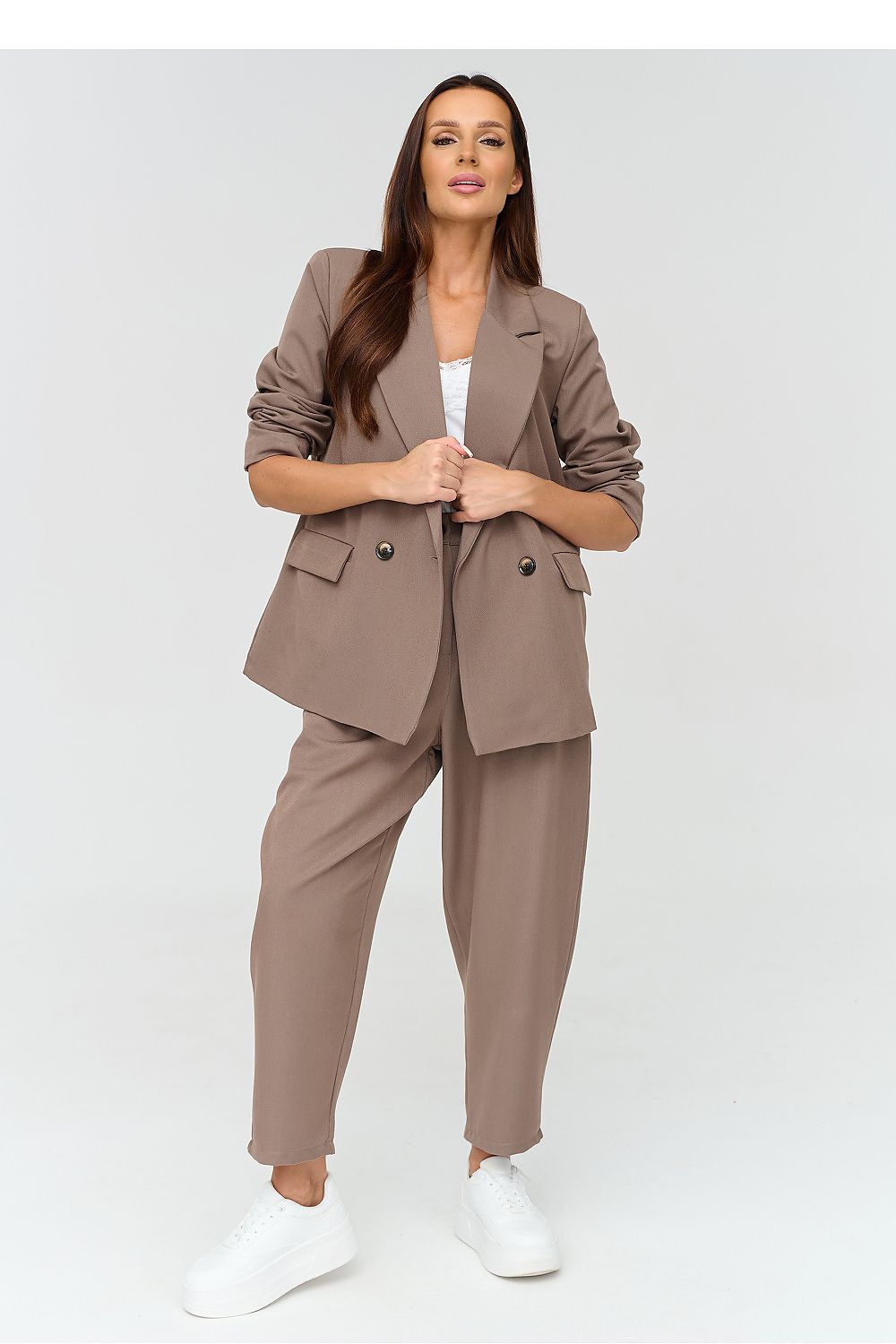 A woman modeling a taupe brown two-piece blazer and trouser set with a white top and white sneakers, shot against a light gray studio background.
