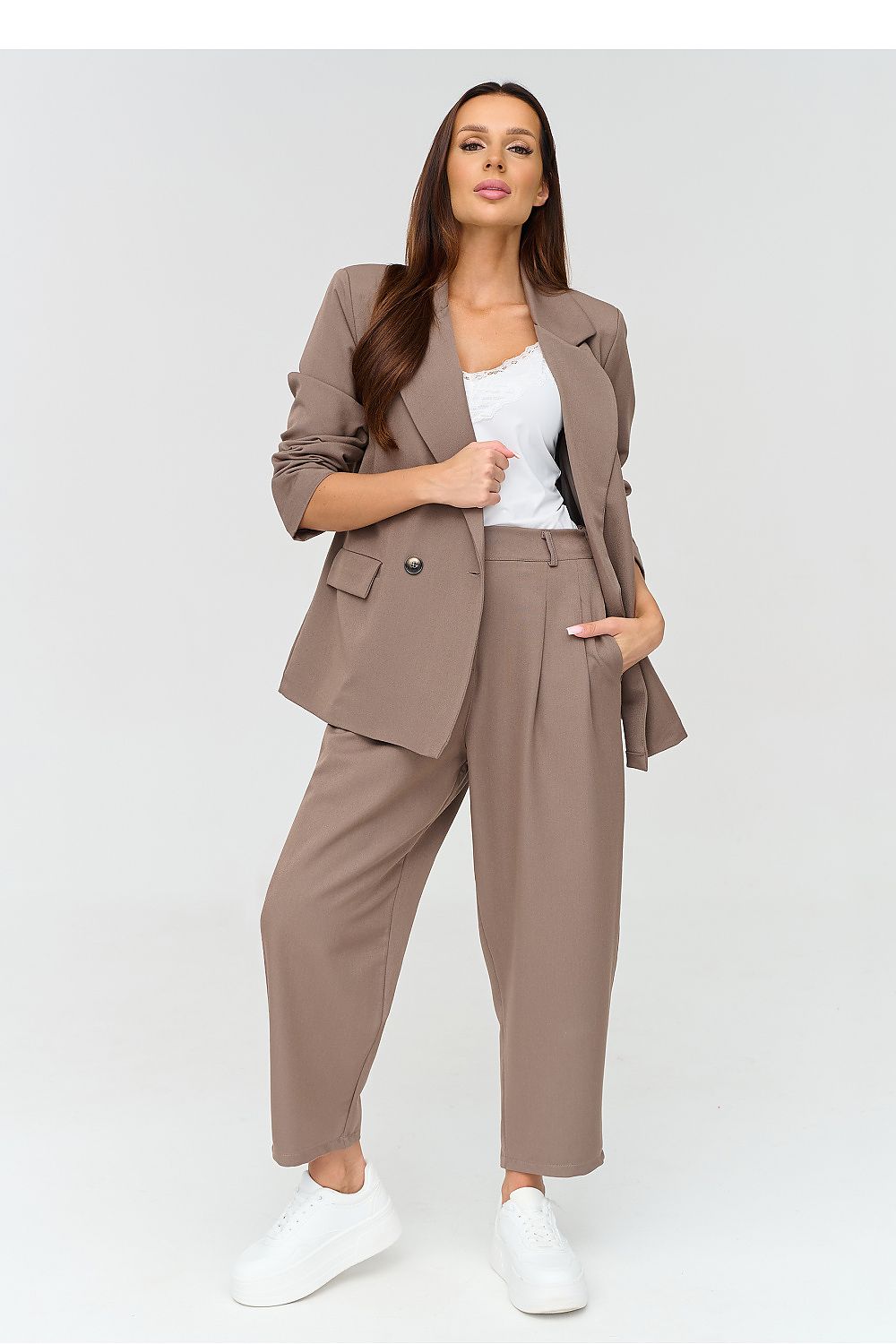 A woman modeling a taupe brown two-piece blazer and trouser set with a white tank top and white sneakers, shot against a light gray studio background.