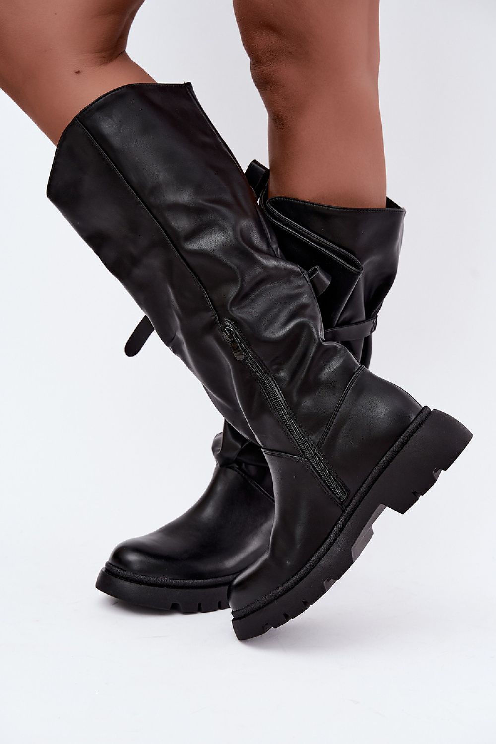 A pair of black knee-high leather boots with side zippers and buckle straps, shown on a model's legs against a white studio background.