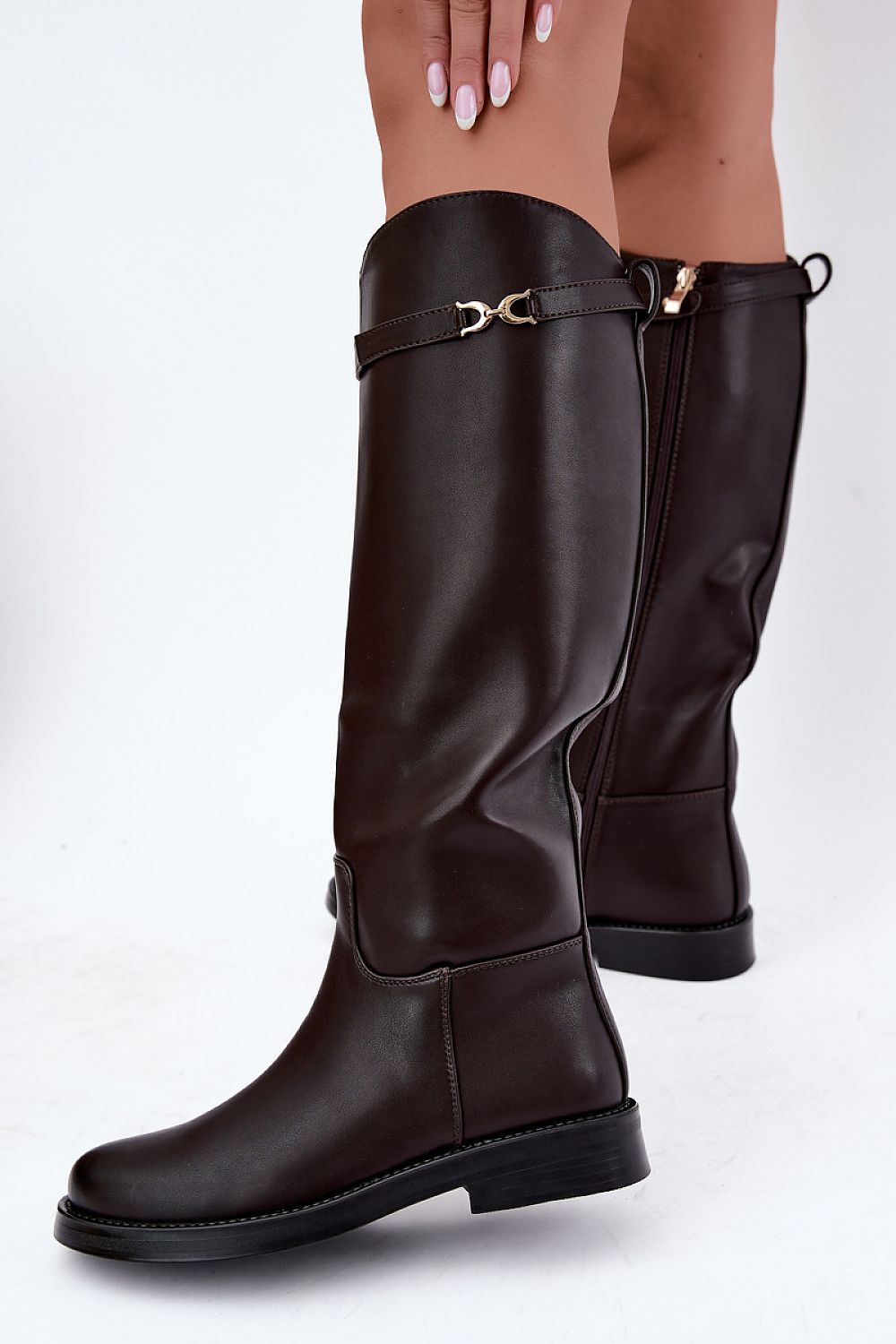 Dark brown knee-high leather boots with a decorative strap and gold buckle detail on the upper shaft, shown on a model against a white studio background.