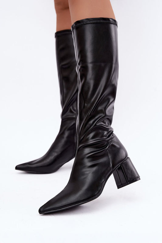 A pair of black leather knee-high boots with pointed toes and block heels, shown on a model's legs against a plain white studio background.