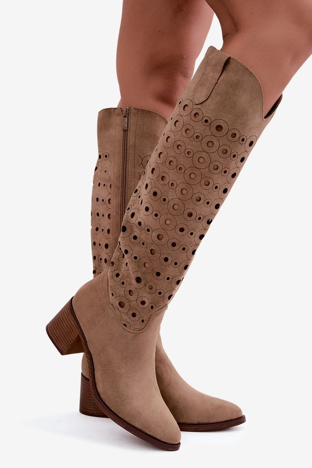 A pair of knee-high taupe suede cowboy boots with circular cutout detailing on the shaft, shown on a model against a plain white background.