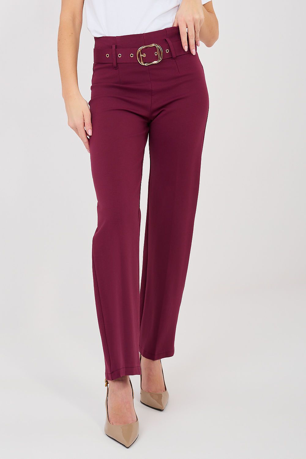 A model wearing a pair of high-waisted, straight-leg trousers in deep burgundy with a matching belt and gold-tone buckle, styled with beige heels against a light gray studio background.