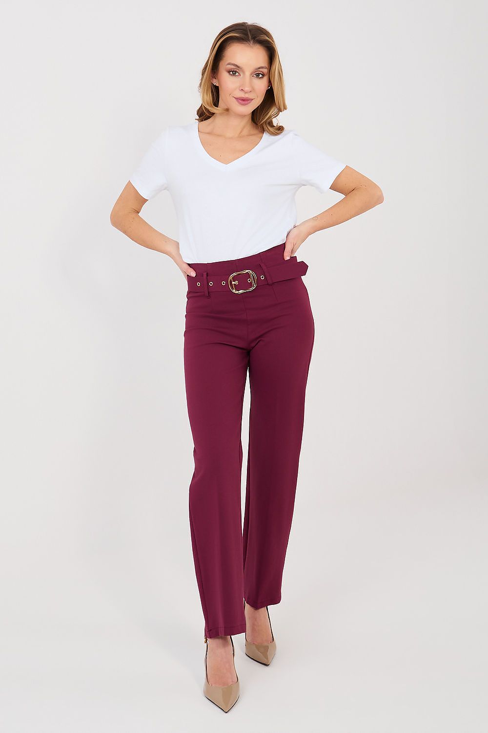 A woman modeling a pair of high-waisted, straight-leg trousers in deep burgundy with a matching belt and gold buckle, paired with a white t-shirt and beige heels, against a light gray studio background.