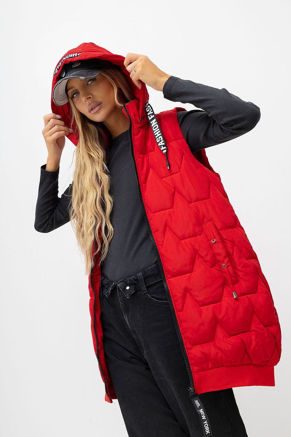 A woman models a bright red hooded puffer vest over a black long-sleeve shirt and black jeans against a plain light gray studio background.