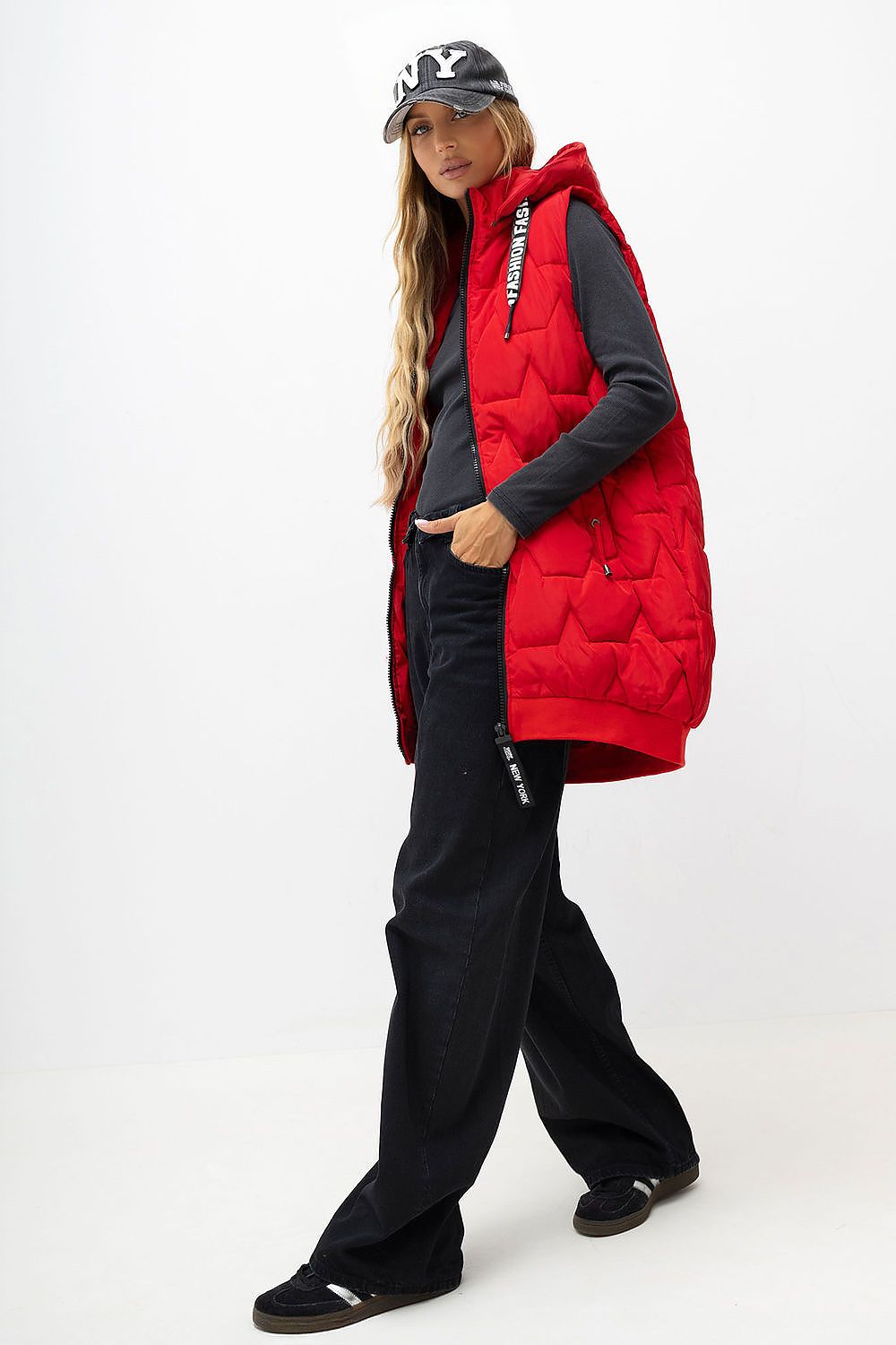 A woman modeling a bright red quilted sleeveless puffer vest over a black long-sleeve top and black wide-leg pants, wearing a black baseball cap, against a plain white studio background.