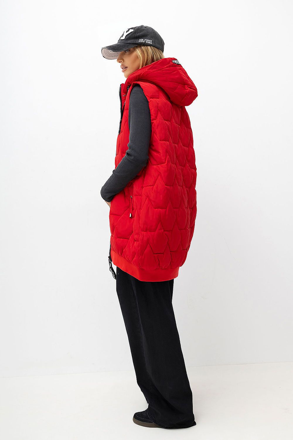 A woman shown in profile wearing a bright red quilted puffer vest over a black long-sleeve top and black pants, standing against a clean white studio background.