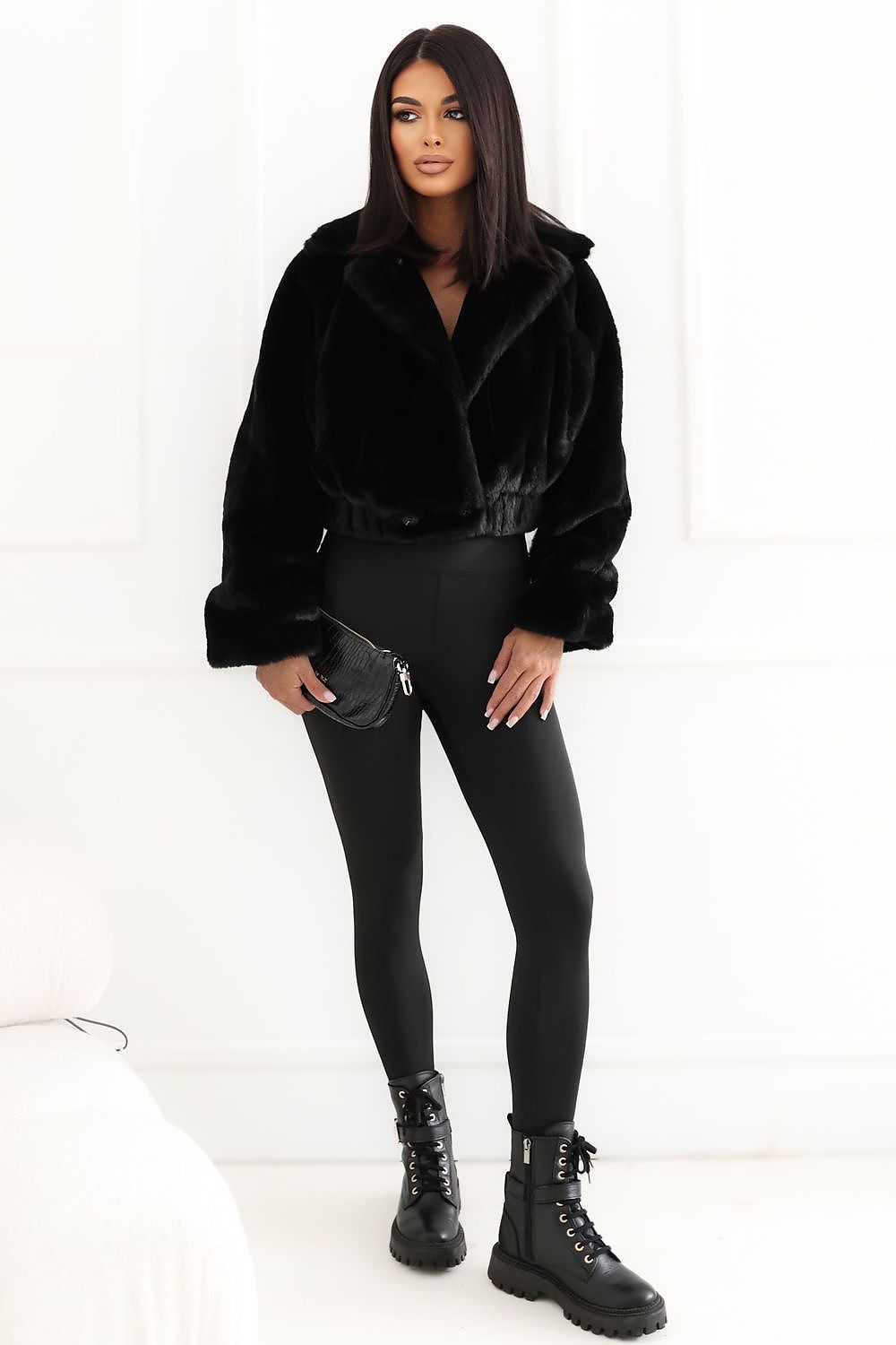 A woman modeling a black faux fur cropped jacket, black leggings, and black combat boots, holding a small black clutch bag against a white paneled wall background.