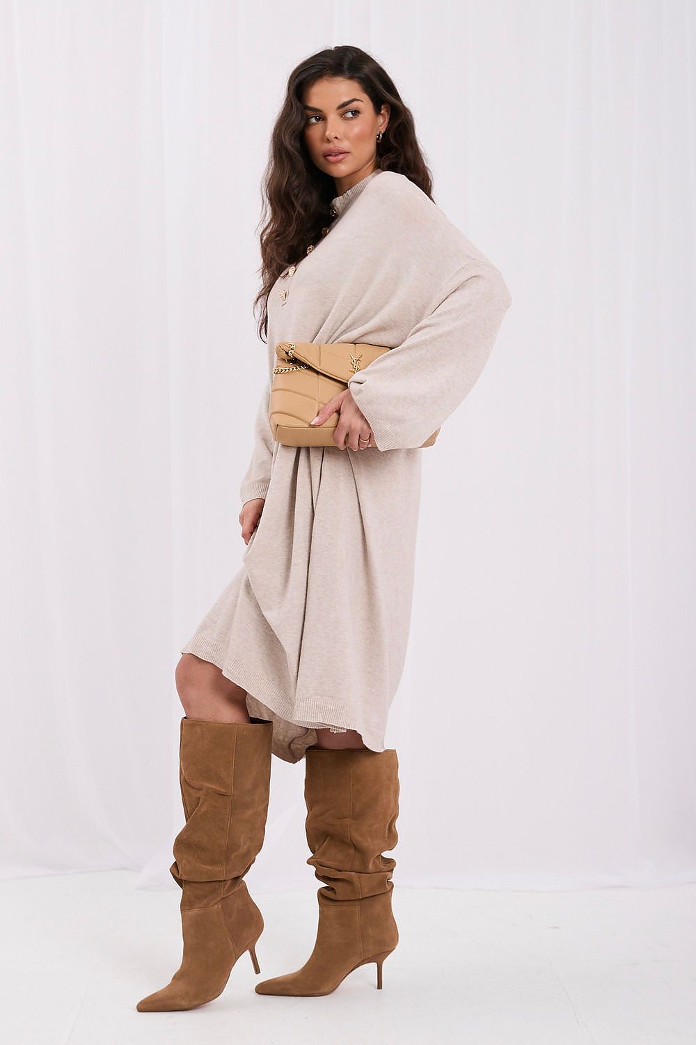 A woman modeling a light beige oversized shawl or wrap dress with wide sleeves, paired with brown knee-high slouchy boots and a tan structured handbag, against a plain white studio background.