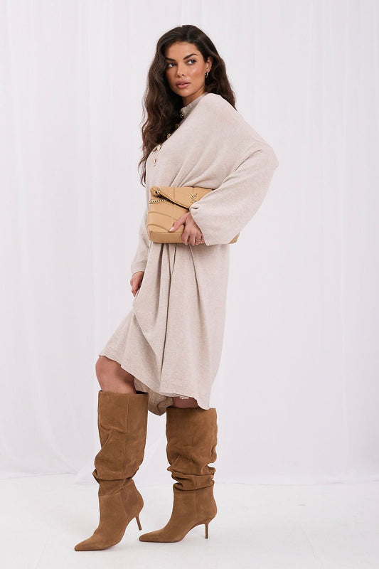A woman modeling a light beige oversized shawl or wrap dress with wide sleeves, paired with brown knee-high slouchy boots and a tan structured handbag, against a plain white studio background.