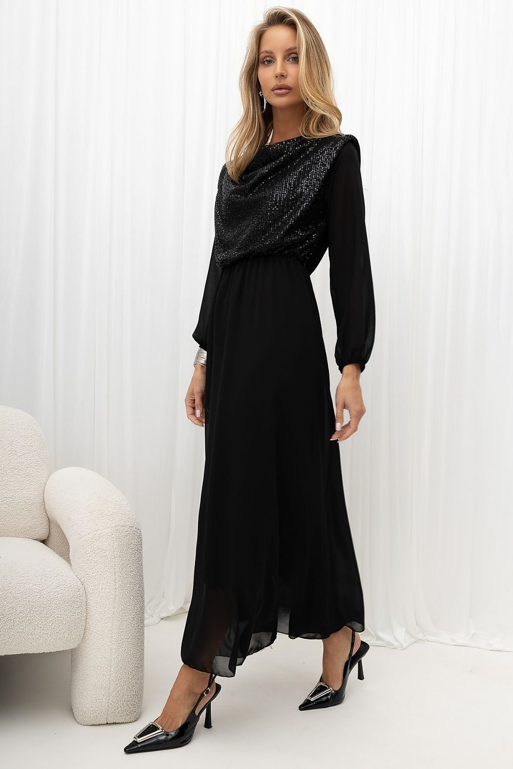 A woman models a long black dress with a textured, sequined cowl neckline and long sleeves, paired with black pointed-toe heels, set against a white curtain backdrop.