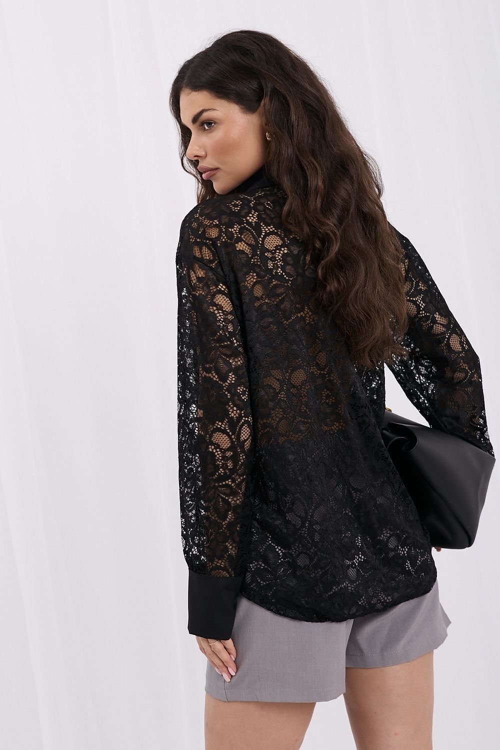 A woman wearing a black lace long-sleeve blouse with a high neck, paired with gray shorts and carrying a black shoulder bag, photographed against a white studio backdrop.