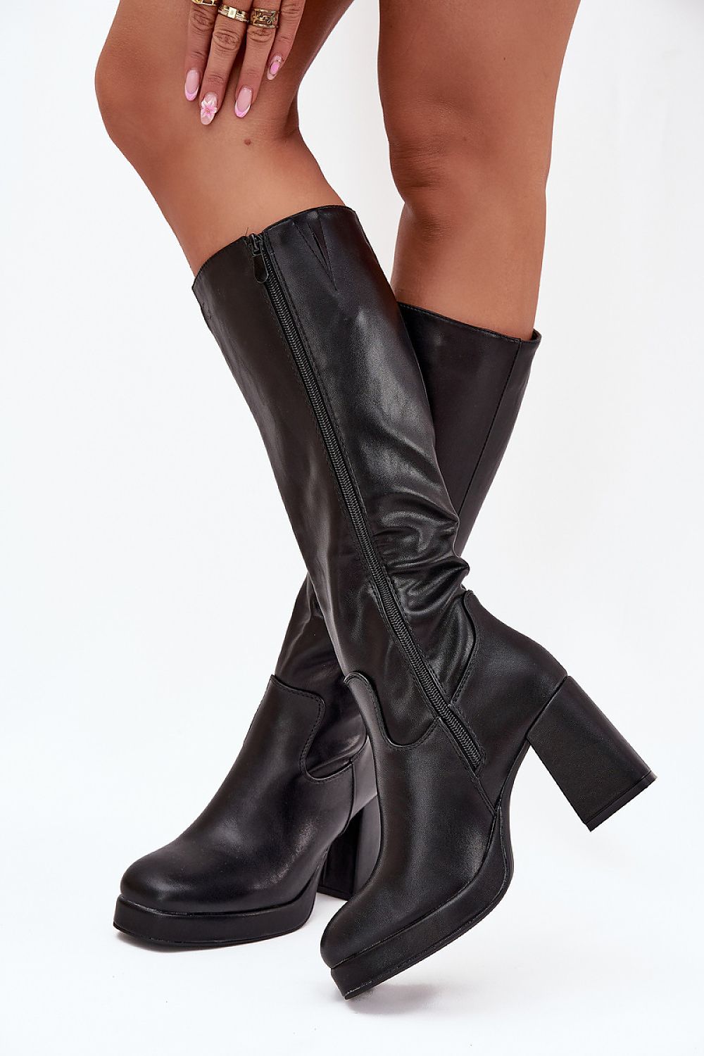 A pair of black knee-high platform boots with a block heel, worn by a model against a plain white studio background.