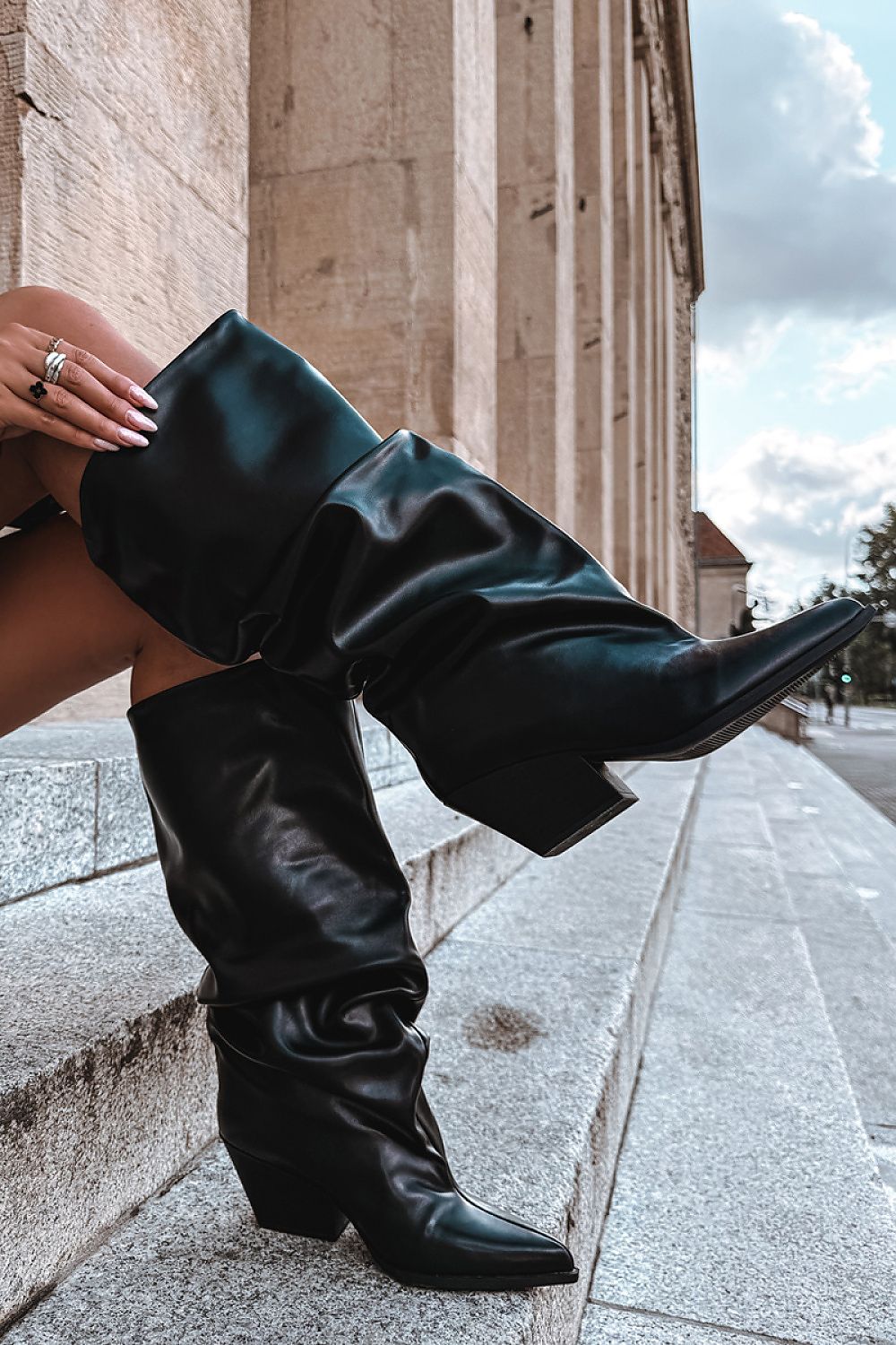 A person seated on stone steps wearing a pair of black knee-high leather boots with pointed toes and low block heels, photographed outdoors beside a stone building.