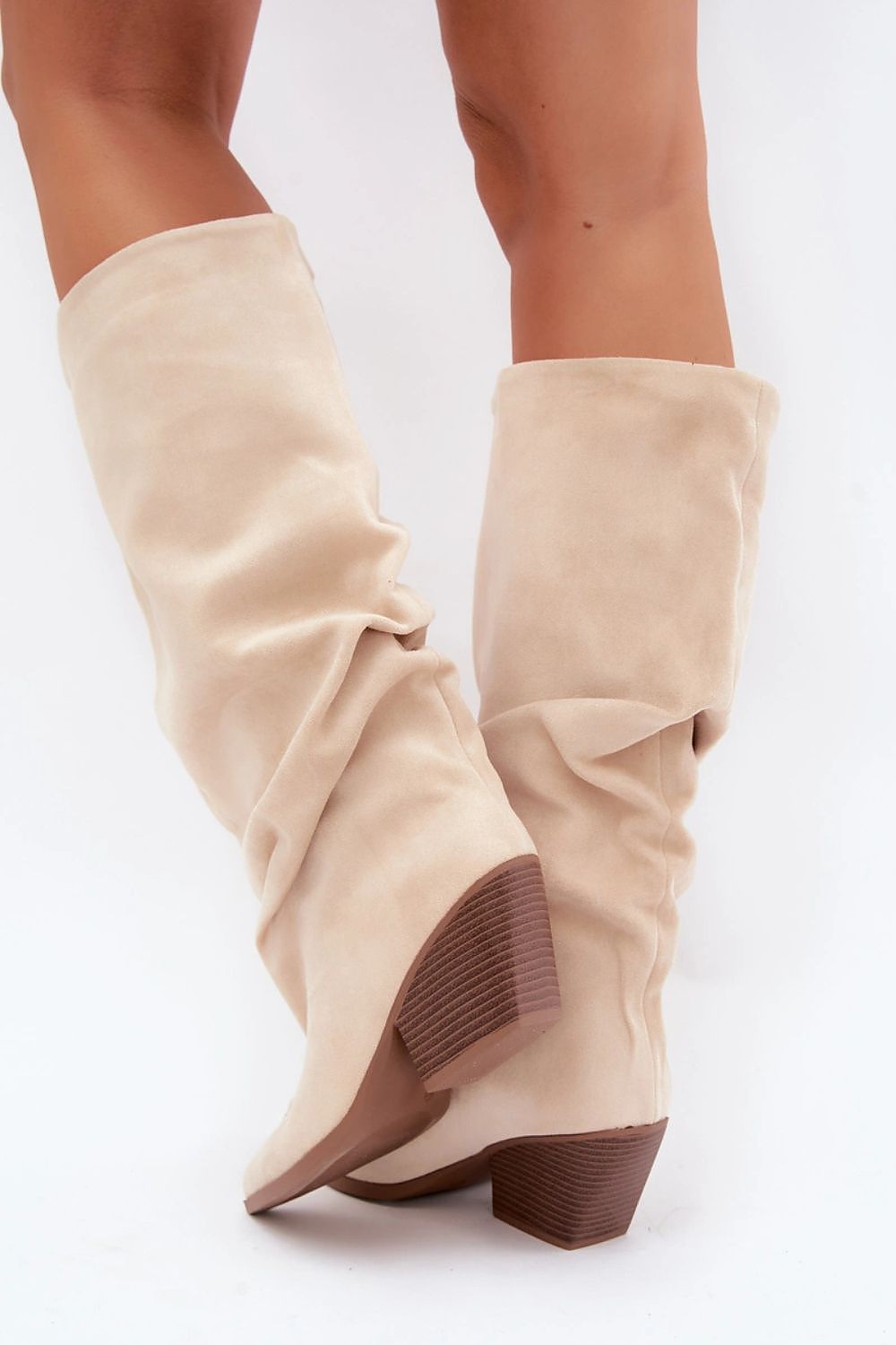 A pair of knee-high slouchy boots in a light beige suede-like material with a brown stacked block heel, shown on a model's feet against a white background.