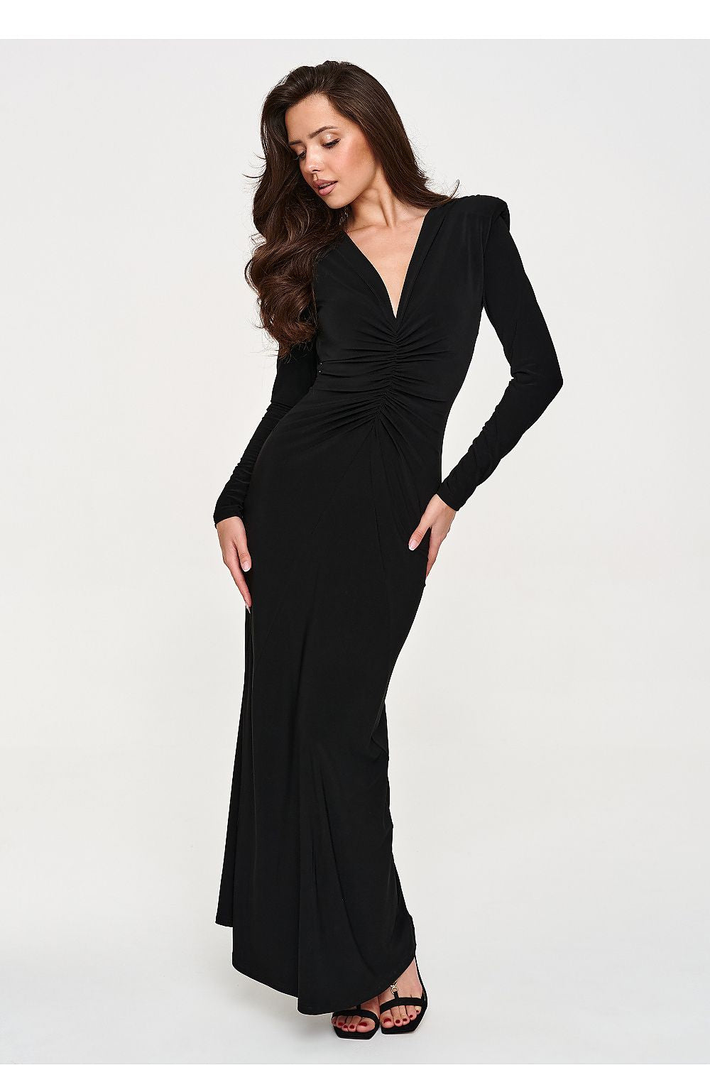 A woman modeling a long-sleeve black maxi dress with a deep V-neckline and gathered ruching at the waist, photographed against a plain light gray studio background.