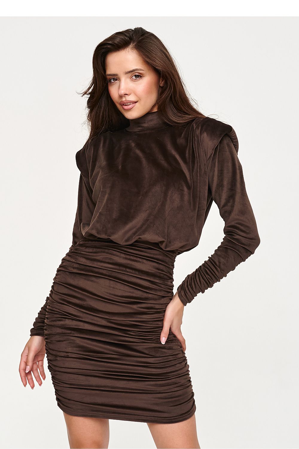 A woman modeling a dark brown velvet long-sleeve dress with a mock neck and ruched skirt, set against a plain light gray background.