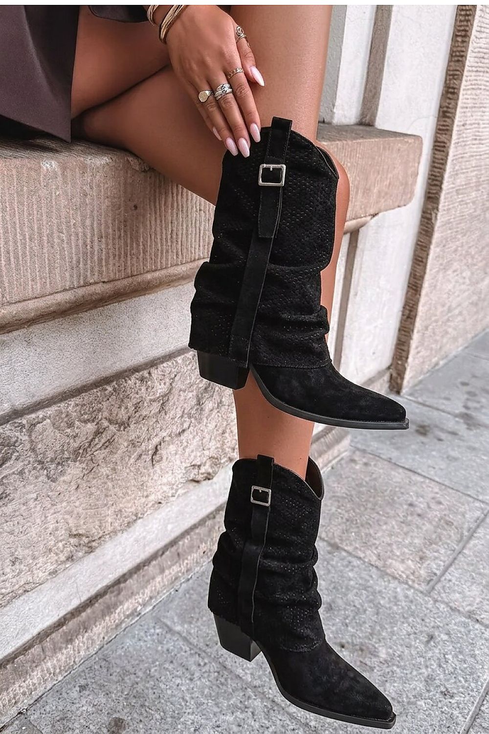 A person wearing black ankle boots with a textured finish, draped side straps, and silver buckle details, sitting on stone steps outdoors.