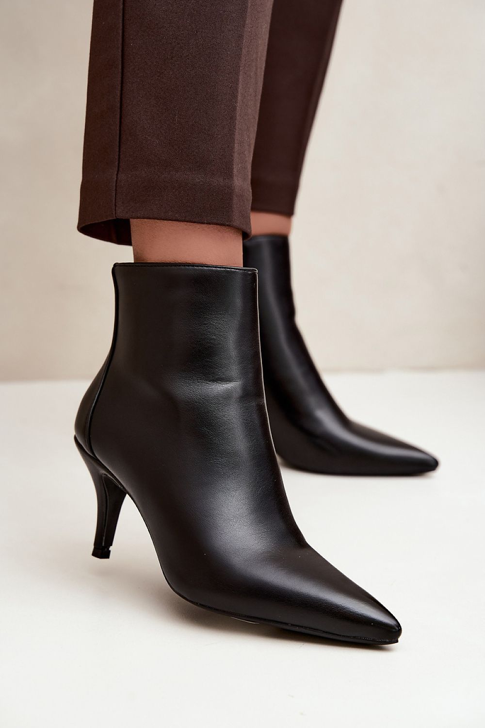 Black pointed-toe ankle boots with kitten heels, shown on a model wearing brown trousers against a neutral beige background.