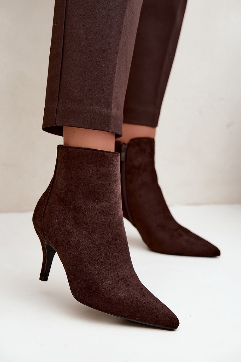 Dark brown suede ankle boots with pointed toes and kitten heels, worn with matching dark brown trousers against a light beige background.