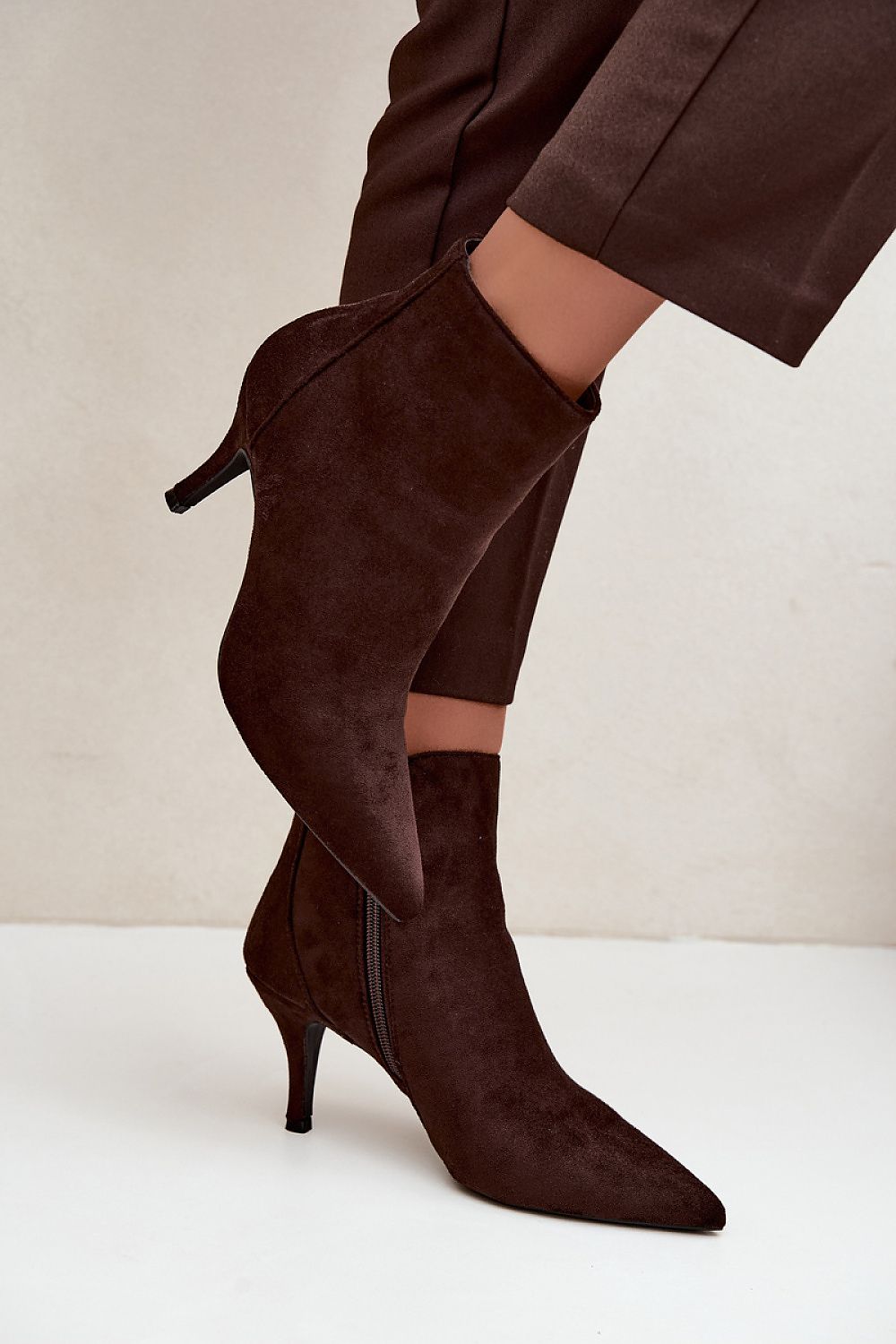 Dark brown suede pointed-toe ankle boots with kitten heels, shown on a model wearing matching brown trousers against a neutral off-white background.
