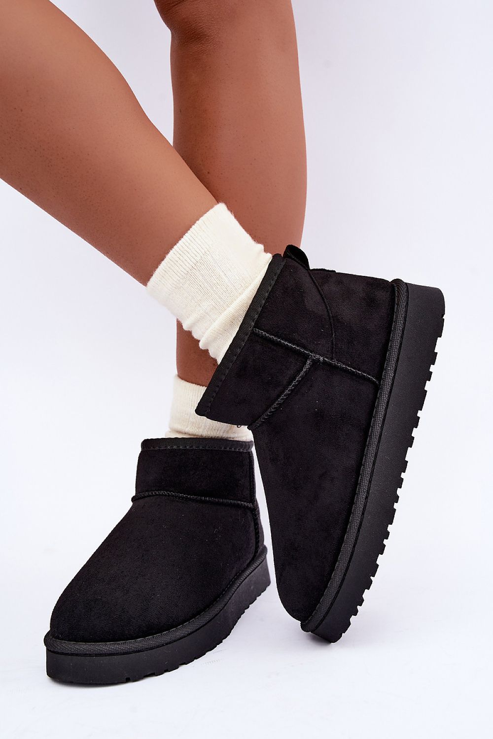 A person wearing black suede ankle boots with thick black soles and white socks, shown from the calves down against a plain white studio background.