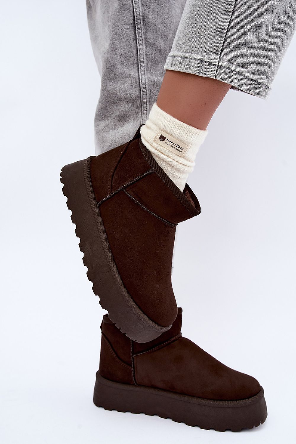 A person wearing dark brown suede platform ankle boots with chunky lug soles, paired with light gray jeans and a cream crew sock.