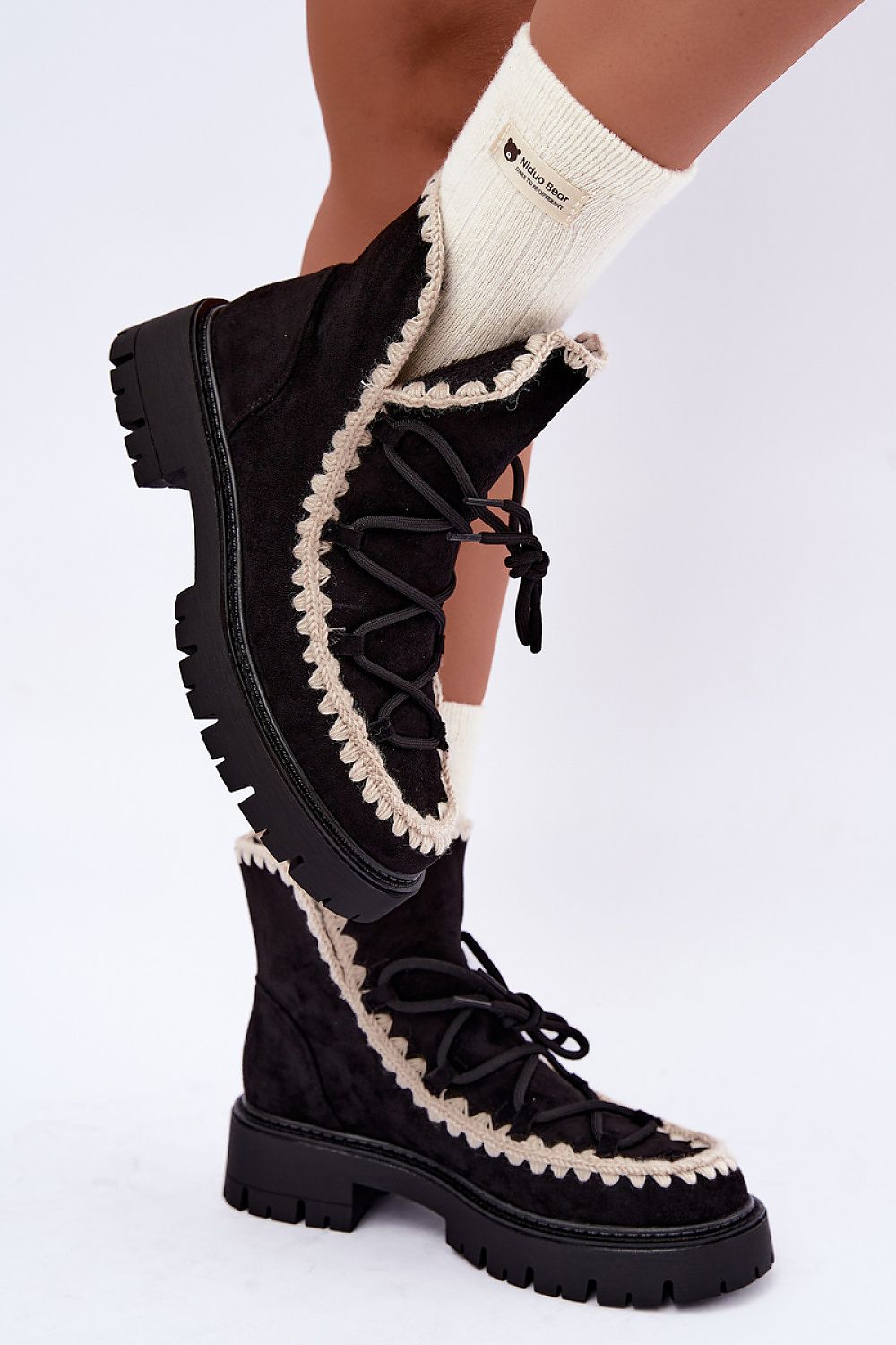 Black suede lace-up ankle boots with a cream-colored fringed trim, shown on a model's feet with cream socks against a white studio background.