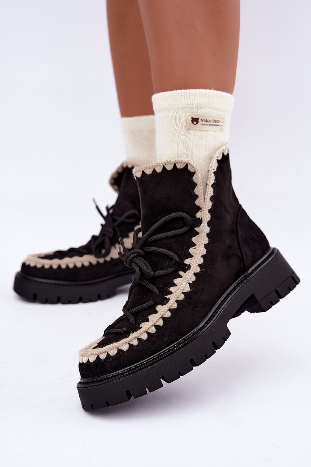 Black suede lace-up ankle boots with cream-colored knit trim and thick black lug soles, shown on a model wearing cream socks against a white studio background.