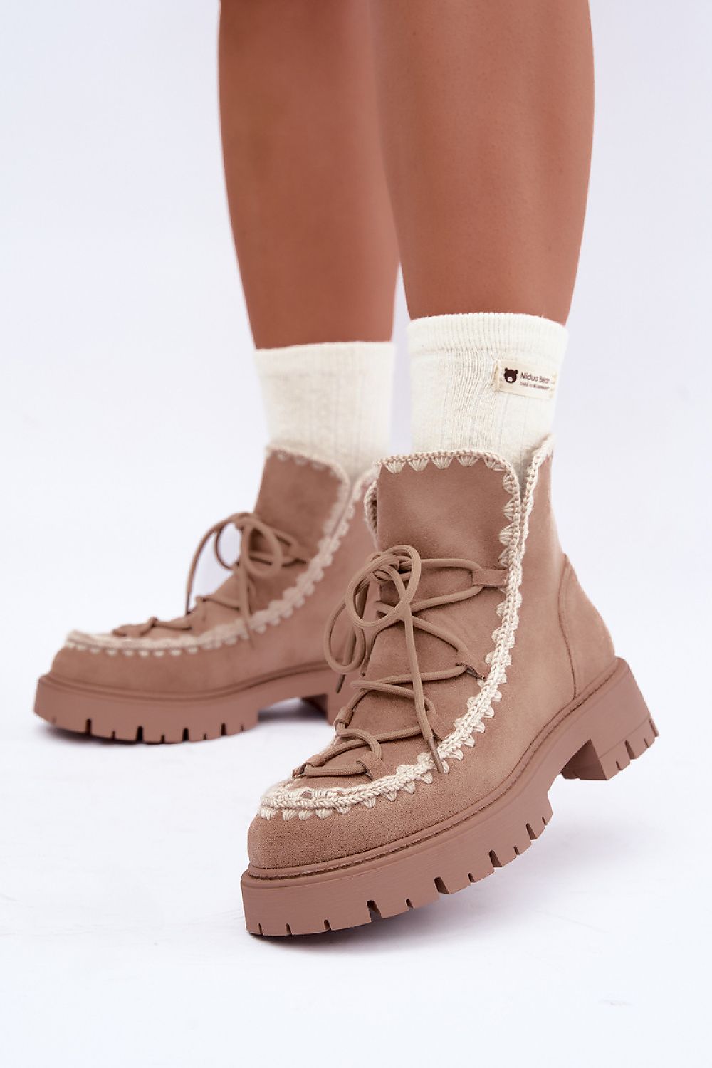 A pair of chunky platform ankle boots in a warm taupe-brown suede-like material, featuring cream-colored decorative trim and laces, shown on a model's feet with cream socks against a white studio background.